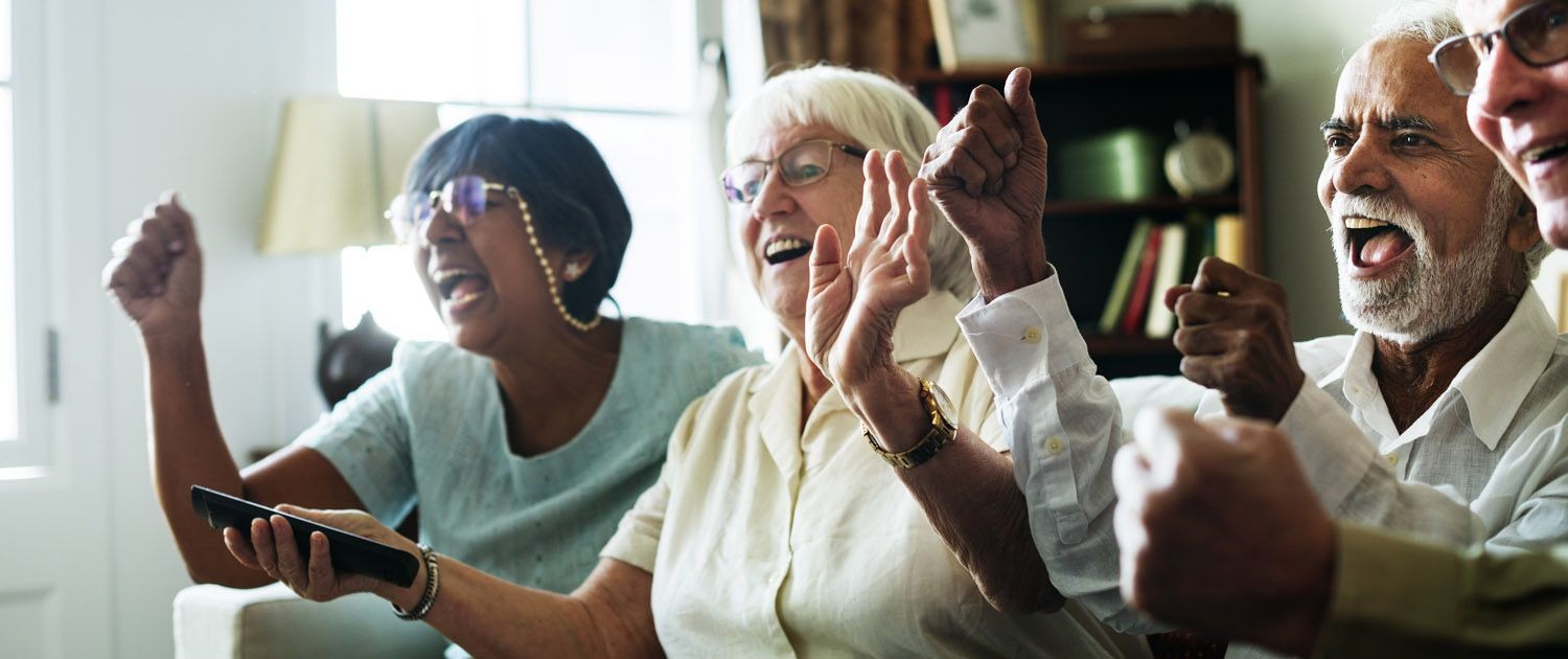 Group of older adults cheering and smiling while watching TV together in a living room.